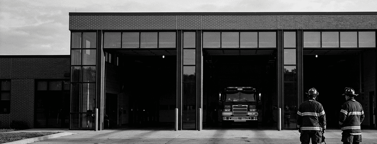 Two firefighters on the apron of a modern fire station at dawn, with an engine parked in the bay